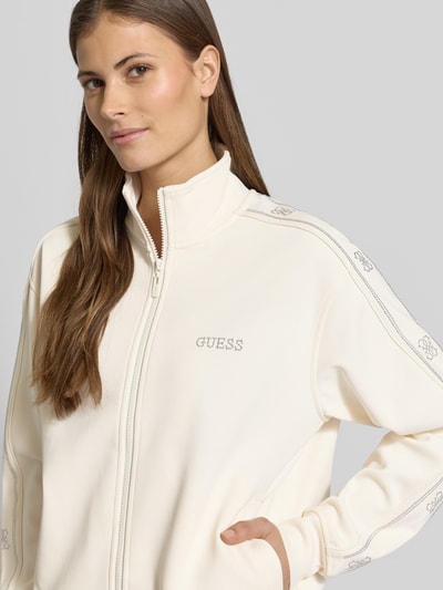 Frauenmodel trägt Regular Fit Sweatjacke mit Viskose-Anteil Modell 'Octavia' von Guess in Creme mit Ziersteinbesatz, Nahaufnahme Oberkörper.