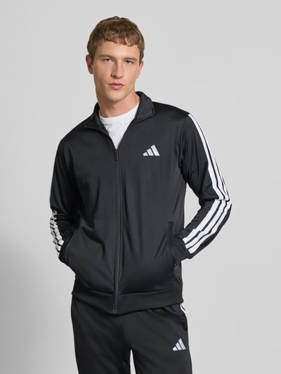 Męski model nosi Trainingsanzug mit Stretch-Anteil od adidas Sportswear, czarny z białymi paskami, elastyczna mieszanka materiałów, prosty krój, stójka, zamek błyskawiczny, elastyczne wykończenia, elastyczny pas, wpuszczane kieszenie, detale z logo, Szerokość w rozmiarze M: 39 cm, długość tyłu: 70 cm, szerokość ramion: 44 cm, długość rękawów: 65 cm, długość wewnętrzna nogawki: 73 cm; całkowity widok z przodu