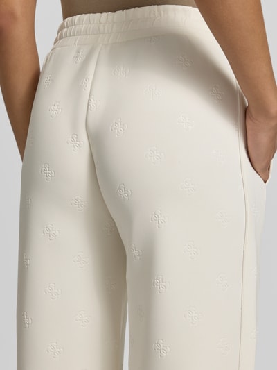 Frauenmodel trägt Wide Fit Sweatpants mit Allover-Logo-Print Modell 'Calista' von Guess Activewear, cremefarben, elastisches Material, Detailaufnahme Rückansicht.