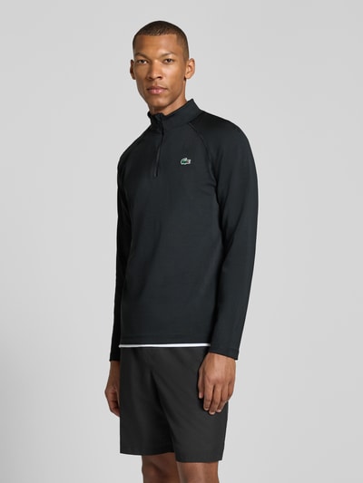 Męski model nosi Regular Fit sportową bluzkę z długim rękawem z technologią ultra-dry™ od Lacoste, kolor czarny, widok z przodu całej sylwetki. Koszulka sportowa Lacoste.