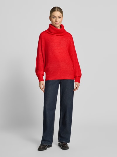 Frauenmodel trägt Relaxed Fit Strickpullover mit Rollkragen Modell 'IHKAMARA' von ICHI in kräftigem Rot, kombiniert mit dunkler Hose, Material-Mix mit Alpaka-Anteil, elastische Abschlüsse, Ganzkörperansicht von vorne.