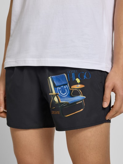 Mannenmodel draagt Badeshorts met motiefprint van Hugo Blue in effen kleuren look met motiefapplicatie, popeline materiaalverwerking, elastische boord, tunnelkoordsluiting, achterzak en zij-steekzakken, detailopname bovenbenen.