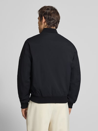Męski model nosi bomberkę z zamkiem błyskawicznym i wpuszczanymi kieszeniami Bomberjacke mit Reißverschluss und Eingrifftaschen von JACK & JONES premium, krój regular fit, stójka, czarny materiał o gładkiej fakturze, długość tyłu w rozmiarze M: 66 cm, szerokość ramion w rozmiarze M: 42 cm, szerokość w rozmiarze M: 60 cm, długość rękawów w rozmiarze M: 68 cm, widok całej sylwetki od tyłu