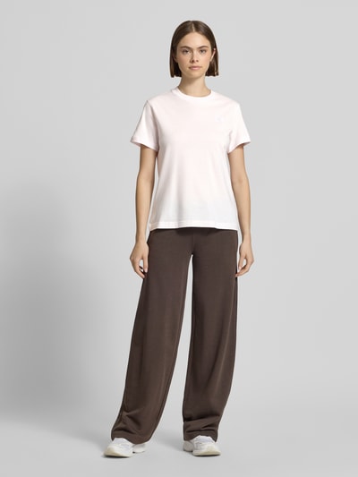 Frauenmodel trägt T-Shirt mit Label-Stitching und Rundhalsausschnitt von New Balance aus Reine Baumwolle im Regular-Fit, Ganzkörperansicht von vorne. Rückenlänge bei Größe S: 56 cm, Farbkontrast zwischen hellem T-Shirt und dunkler Hose, glatte Stofftextur.