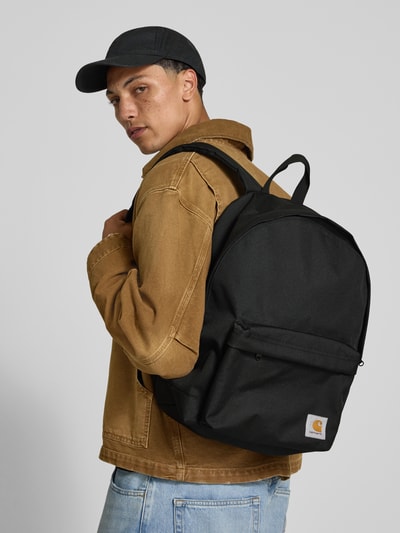 Männermodel trägt zwarte rugzakfunctie, labelpatch, buitenvak met ritssluiting, draagband, verstelbare schouderband, Carhartt Work In Progress Rucksack mit Label-Patch Modell 'Jake', hoofvak met tweewegritssluiting, mesh binnenvak met ritssluiting, breedte 40 cm, lengte 45 cm, diepte 14 cm, schouderbandlengte 68 cm, Ganzkörperansicht van voren.