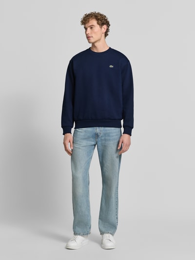 Männermodel trägt Regular Fit Sweatshirt aus Baumwoll-Mix von Lacoste in Dunkelblau und Jeans, Ganzkörperansicht von vorne. Herren Sweatshirt von Lacoste