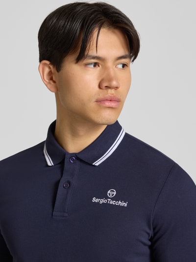 Męski model nosi Slim Fit Poloshirt mit Logo-Stitching Modell 'Reed' od Sergio Tacchini, granatowa, elastyczna bawełna, krój slim fit, krótki rękaw, kołnierzyk polo, szczegóły haftu logo, kontekst: długość tyłu w rozmiarze M: 71 cm, zbliżenie na górną część ciała.