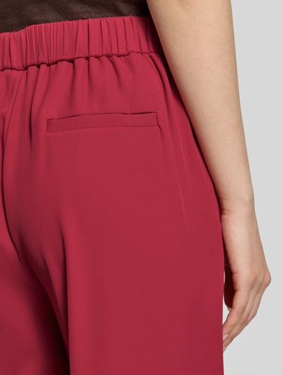 Kobieta nosi spodnie materiałowe 'Stoffhose mit elastischem Bund und Eingrifftaschen' od Jake*s Collection, mieszanka materiałów z dodatkiem streczu, krój wide leg fit, elastyczny pas, wpuszczane kieszenie, z tyłu kieszeń z wypustką, szerokość w rozmiarze 36: 38 cm, długość wewnętrzna nogawki w rozmiarze 36: 78 cm, widoczna faktura gładka, kolor ciemnoczerwony, detal zdjęcia tylnej części spodni, zbliżenie na pas i kieszeń, detalaufnahme.