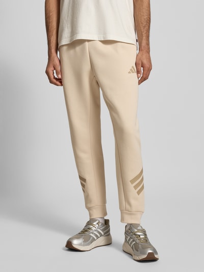 Männermodel trägt Regular Fit Sweathose mit Logoprint von adidas Sportswear, katoenmix, elastische band met tunnelkoord, regular fit, elastische beenboorden, binnenbeenlengte maat M 71 cm, beige en goud tinten, gladde textuur. Nahaufnahme onderlichaam, voorkant.