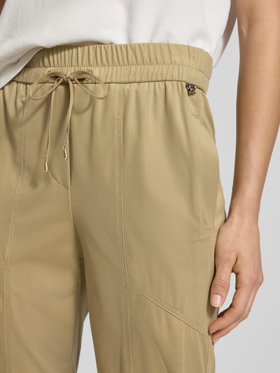 Kobiece modelka nosi Slim Fit Stoffhose mit Reißverschlusstaschen von Marc Cain w beżowym kolorze, elastyczny materiał, krój slim fit, elastyczny pas z troczkiem, detale z logo, kieszenie na zamek; Szerokość w rozmiarze 36: 37 cm, długość wewnętrzna nogawki: 68 cm, zbliżenie na górną część ciała
