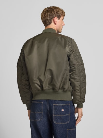 Männermodel trägt Bomberjacke mit Pattentaschen und Label-Applikation von Alpha Industries in Olivgrün, glatte Textur, Ganzkörperansicht Rückansicht, Regular Fit, Stehkragen, Reißverschluss, Kontext: Rückenlänge 69 cm, Schulterbreite 44 cm, Brustweite 54 cm, Ärmellänge 64 cm.