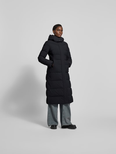 Vrouwenmodel draagt Daunenmantel mit Kapuze van Moose Knuckles in zwart, gewatteerd, lange lengte, taillering, kap met rits, zakken, stretchmateriaal. Volledige lichaamsaanzicht van voren.