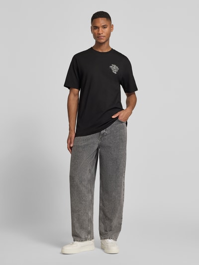 Męski model nosi T-Shirt mit rückseitigem Motiv-Print Modell 'Ben Faience' von Les Deux, czarny kolor, prosta bawełna, krój regular fit, okrągły dekolt, nadruk na piersi, widoczny detal marki, długość tyłu w rozmiarze M: 70 cm, widok całej sylwetki z przodu.