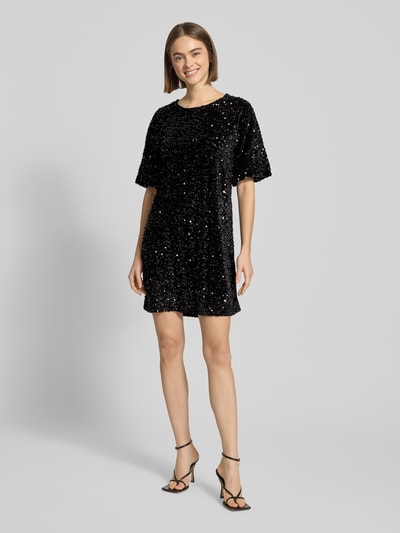 Kobiece model prezentuje sukienkę koktajlową z cekinowym obszyciem Schmales Cocktailkleid mit Paillettenbesatz Modell 'KAM' od Pieces, efekt: jednolity kolor, efekt błyszczący, krój wąski, długość mini, dekolt w łódkę, wyhaftowane logo, aksamitna powierzchnia, krótki rękaw, prosty dół, całość czarna, widok całej sylwetki z przodu.