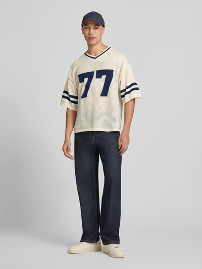 Mężczyzna nosi Strick-Trikot mit Jacquard Muster von REVIEW w odcieniach kremowym i granatowym, perforowanie, prosty krój, dekolt w serek, rękaw o dł. 1/2, długość tyłu w rozmiarze M: 69 cm, trykotowy styl, Ganzkörperansicht von vorne.