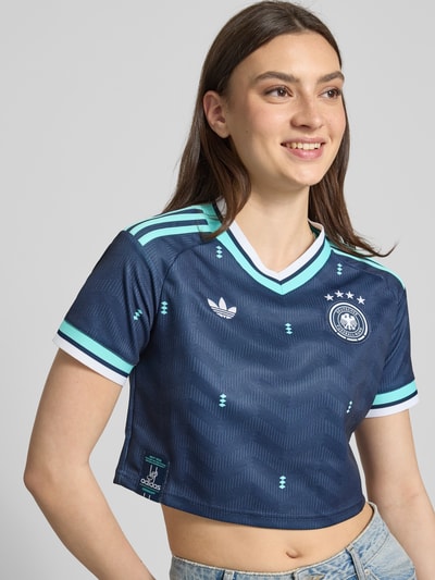 Frauenmodel trägt DFB Cropped Auswärtstrikot 2026 von adidas Sportswear, mehrfarbig mit V-Ausschnitt, Logo und Wappen, Regular Fit, Nahaufnahme Oberkörper.