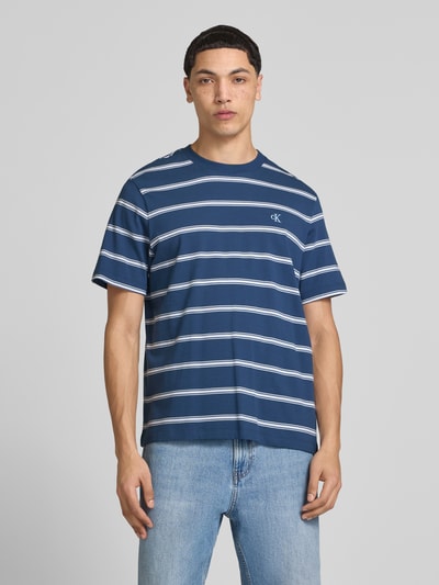 Männermodel trägt T-Shirt mit Logo-Stitching von Calvin Klein Jeans in Blau mit weißen Streifen, Reine Baumwolle, Gerader-Schnitt, Rundhalsausschnitt, Rückenlänge bei Größe M: 69 cm, Ganzkörperansicht von vorne.