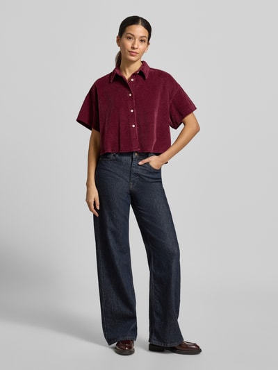 Frauenmodel trägt Cropped Hemdbluse mit Knopfleiste Modell 'PADOW' von American Vintage in dunklem Rot, reine Baumwolle, lockerer Schnitt, Hemdblusenkragen, halbe Ärmel, dazu dunkelblaue Jeans, Ganzkörperansicht von vorne.