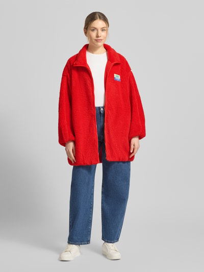 Frauenmodel trägt Oversized Teddyjacke mit Stehkragen Modell 'HOKTOWN' von American Vintage in leuchtend Rot, Reißverschluss, strukturierte Oberfläche, Eingrifftaschen, Ganzkörperansicht von vorne, Kontext: Damen Teddyjacke oversized geschnitten, Label-Detail, Rückenlänge 89 cm, Schulterbreite 58 cm, Brustweite 67 cm, Ärmellänge 49 cm.