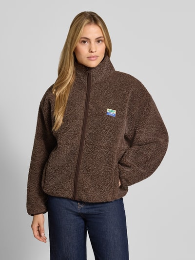 Frauenmodel trägt Regular Fit Jacke aus Teddyfell Modell 'Hoktown' von American Vintage in Dunkelbraun, Logo-Patch, Stehkragen, Eingrifftaschen, Reißverschluss, strukturierte Oberfläche, Ganzkörperansicht von vorne.