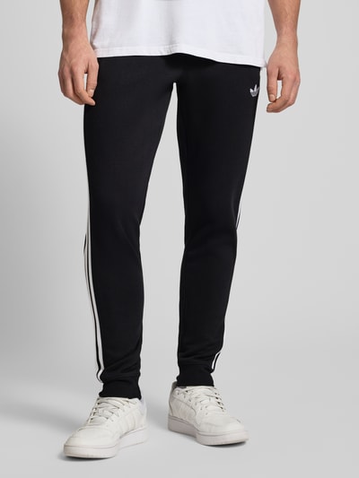 Männermodel trägt Sweatpants mit elastischem Bund von adidas Originals in Schwarz mit weißen Streifen, Material-Mix mit Stretch-Anteil, lockerem Schnitt, Label-Stitching und französischen Eingrifftaschen, Ganzkörperansicht von vorne.