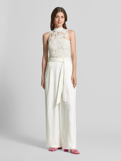 Frauenmodel trägt Jumpsuit mit Häkelspitze Modell 'CALANTA' von Ted Baker aus weißem Baumwoll-Mix, Taillierter Schnitt, Bindegürtel, Seitliche Eingrifftaschen, gerippter Rundhalsausschnitt, Ganzkörperansicht von vorne.