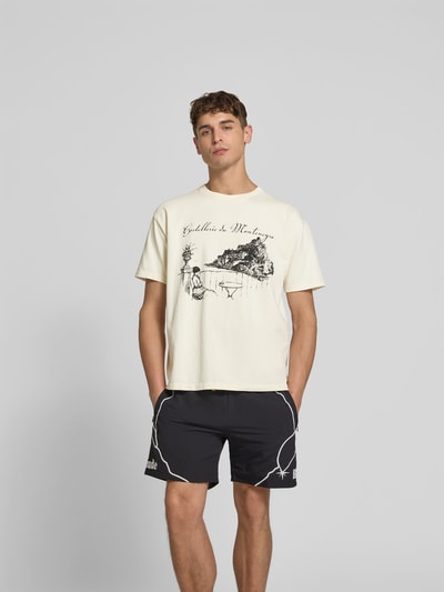 Männermodel trägt Regular Fit T-Shirt mit Motiv-Print von RHUDE aus reiner Baumwolle, Rundhalsausschnitt, heller Stoff, Ganzkörperansicht von vorne.