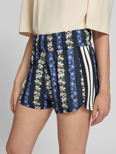 Frauenmodel trägt Regular Fit Sportshorts mit elastischem Bund von adidas, florales Muster in Blau und Weiß, adidas Sportswear, Nahaufnahme Hüfte.
