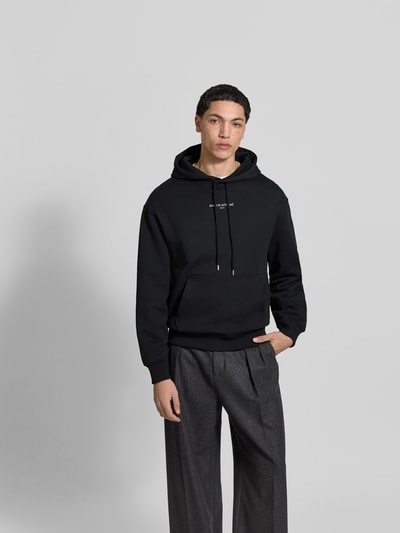 Männermodel trägt Hoodie mit Label-Stitching von MAISON KITSUNÉ in Schwarz, lockere Passform, Känguru-Tasche, Nahaufnahme Oberkörper.