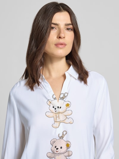 Modelka kobieca nosi Bluse aus Seiden-Mix mit Motiv-Print von HERZENS, biała bluzka o kroju loose fit z mieszanki jedwabiu z dodatkiem streczu, kołnierzyk koszulowy, dekolt w serek, nadruk z motywem misia, wydłużony tył, zaokrąglony dół, długość tyłu 69 cm w rozmiarze 36, długość rękawa 60 cm w rozmiarze 36, tekstura gładka, widoczne detale nadruku, półzbliżenie górnej części ciała.