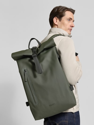 Männermodel trägt Rolltop Rucksack mit Reißverschlusstasche von Rains in Olivgrün, Karabinerhaken, Tragehenkel, verstellbare Träger, Ganzkörperansicht Rückansicht. Breite 31 cm, Länge 61 cm, Tiefe 15 cm, Schulterriemen 74 cm.