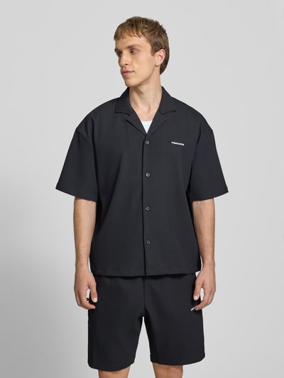 Männermodel trägt Relaxed Fit Freizeithemd mit Reverskragen Modell 'Troy' von Pegador, Schwarz, Strukturmuster, Ganzkörperansicht von vorne. Rückenlänge bei Größe M: 67 cm, Schulterbreite: 49 cm, Ärmellänge: 24 cm, durchgängige Knopfleiste, kurzärmelig, Logo-Stitching.