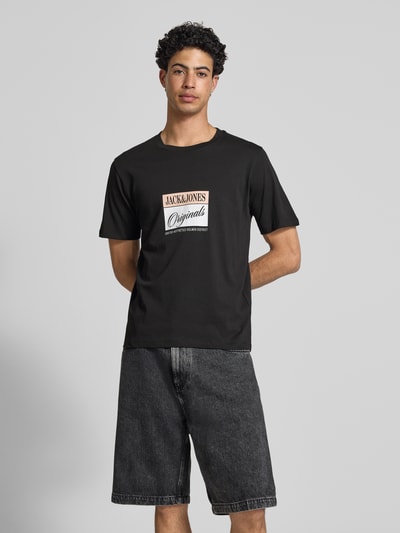 Męski model prezentuje męski t-shirt z nadrukiem z napisem model 'RHYERES' od Jack & Jones z woskowanej bawełny, krój regular fit, okrągły dekolt, czarny z białym i różowym nadrukiem, długość tyłu w rozmiarze M: 67 cm, pełen widok z przodu.