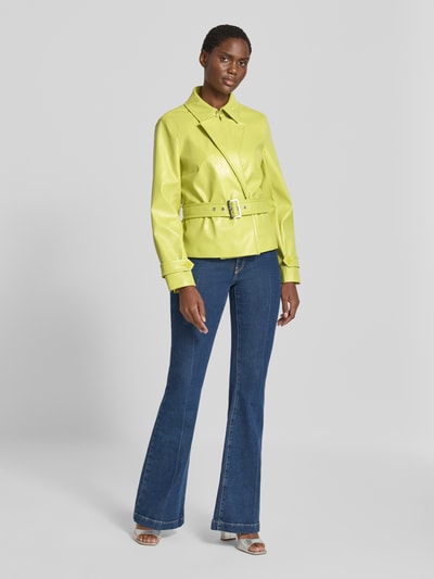 Frauenmodel trägt Jacke mit Gürtel und Ärmelriegeln von Marc Cain in Gelb, sichtbare Struktur, kombiniert mit blauer Jeans, Ganzkörperansicht von vorne, Regular Fit, Hakenverschluss, Rückenlänge 58 cm, Schulterbreite 40 cm, Brustweite 48 cm, Ärmellänge 61 cm.