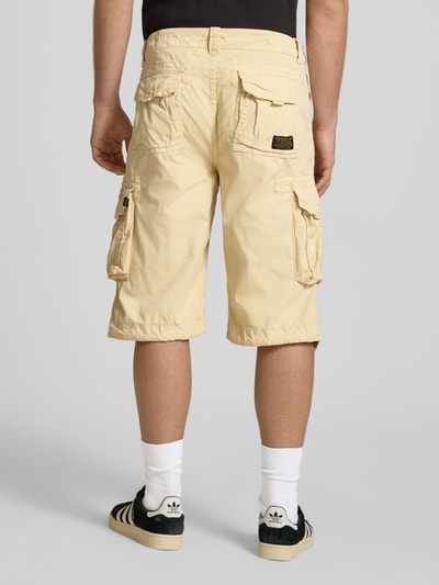 Männermodel trägt Bermudas mit Pattentaschen und Logo-Stitching van Alpha Industries in beige, rechte pasvorm, katoen, zichtbare zakken, ganzkörperansicht rückansicht.