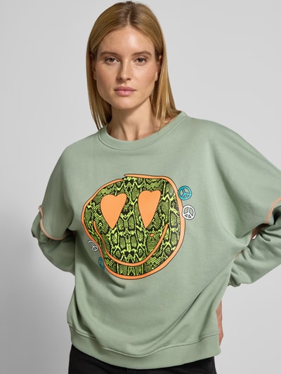 Frauenmodel trägt Oversized Sweatshirt mit Motiv-Print Modell 'Heartface' von miss goodlife in sanftem Grün, reine Baumwolle, elastische Abschlüsse, Ganzkörperansicht von vorne.