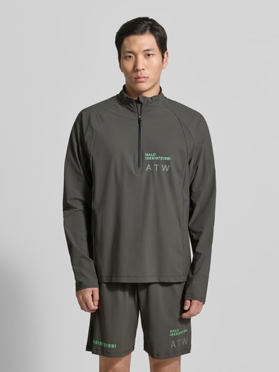 Männermodel trägt Funktionsshirt mit Stehkragen und Reißverschluss Modell 'AIR' von HALO, materiaalmix met stretch, regular fit, labelprint, ruglengte bij maat M 70 cm, donkergrijs met groene details, gladde textuur, ganzkörperansicht van voren.
