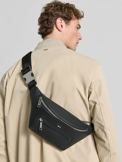 Männermodel trägt Bauchtasche mit Label-Detail Modell 'RAY' von Boss in Optik:Allover-Muster aus Kunstleder, Ganzkörperansicht Rückansicht.