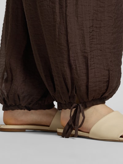 Frauenmodel trägt Ballon Hose aus Viskose-Mix mit Kordelzug am Saum von Gina Tricot, texturierte braune Oberfläche, Detailaufnahme Fußbereich. Damen Balloon HOSE, Loose-Fit, elastischer Bund mit Kordelzug, Saum mit Kordelzug, Bundweite Größe S: 35 cm, Innenbeinlänge Größe S: 70 cm.