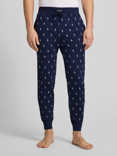 Männermodel trägt Sweathose mit Allover-Logo-Print van Polo Ralph Lauren in donkerblauw met witte logo's, elastische tailleband, Ganzkörperansicht von vorne.