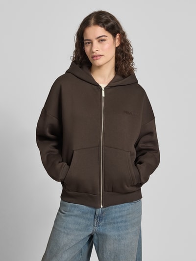 Modelka nosi Sweatjacke mit Logo und Känguru-Tasche von Review w kolorze ciemnego brązu, bawełniany materiał, relaxed fit, kaptur, zamek, kieszeń kangurka, widoczne logo, przód, ujęcie całej sylwetki z przodu.