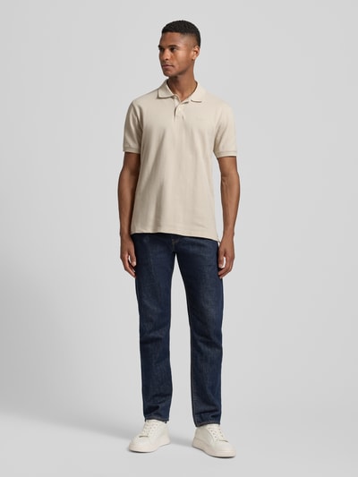 Männermodel trägt Slim Fit Poloshirt mit Label-Stitching von GAP in Beige, kombiniert mit dunkler Jeans, Ganzkörperansicht von vorne.
