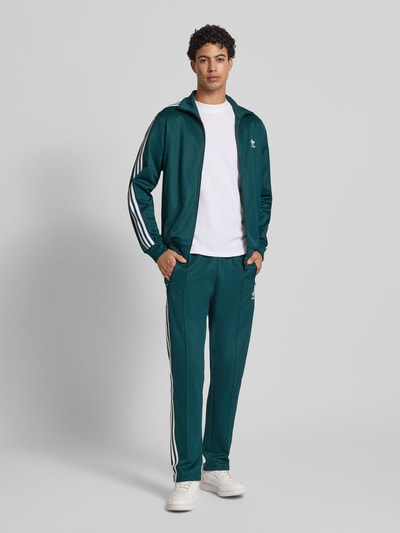 Männermodel trägt Sweatpants mit elastischem Bund von adidas Originals in Dunkelgrün, Baumwoll-Mix, Lockerer-Schnitt, Label-Stitching, Reißverschlusstaschen, Gesäßtasche, Innenbeinlänge 76 cm bei Größe M, Ganzkörperansicht von vorne.