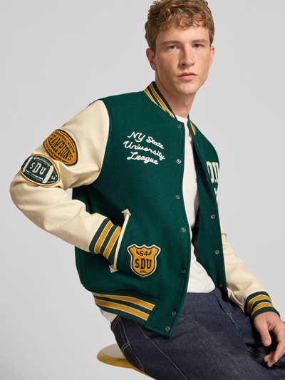 Männermodel trägt College-Jacke aus Woll-Mix mit Eingrifftaschen von Superdry in Grün mit cremefarbenen Ärmeln, gestickten Applikationen, Regular Fit, Stehkragen und Knopfleiste, Nahaufnahme Oberkörper.