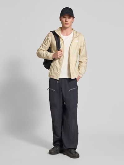 Männermodel trägt Windbreaker mit Logo und ritszakken van The North Face, regular fit, capuchon, ritssluiting, ganzkörperansicht von vorne. Ruglengte bij maat M: 66 cm, schouderbreedte bij maat M: 44 cm, borstwijdte bij maat M: 58 cm, mouwlengte bij maat M: 68 cm. Neutrale tinten crème en zwart, lichte glanzende structuur.