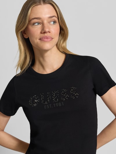 Frauenmodel trägt T-Shirt mit Label-Print und Strasssteinbesatz von Guess, Reine Baumwolle, Straight Fit, Rundhalsausschnitt, Nahaufnahme Oberkörper.