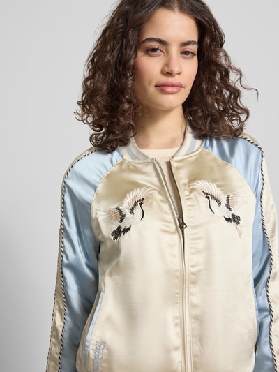 Modelka nosi Bomber Jacke mit Motiv- und Logo-Stitching od Superdry z satyny w kolorach beżowym i jasnoniebieskim, prosty krój, krótki kołnierz, zamek, boczne kieszenie, elastyczne wykończenia, szerokość w rozmiarze S: 53 cm, długość tyłu: 58 cm, szerokość ramion: 45 cm, długość rękawa: 64 cm, zbliżenie na górną część ciała.