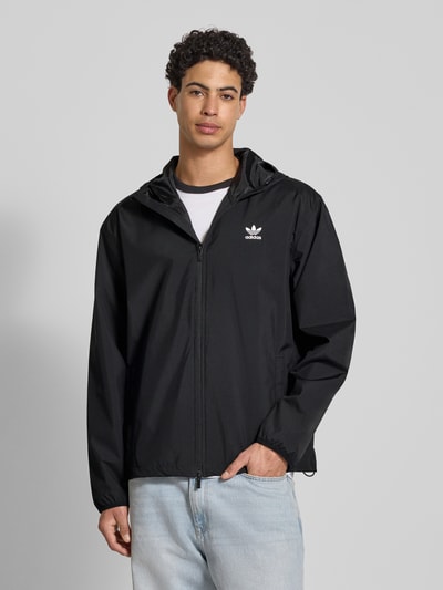 Męski model nosi Kurtka mit Kapuze von adidas Originals, czarna, krój regular fit, kaptur, zamek błyskawiczny, nadruk z logo, wpuszczane kieszenie, długość tyłu w rozmiarze M: 71 cm, szerokość ramion w rozmiarze M: 55 cm, szerokość w rozmiarze M: 61 cm, długość rękawów w rozmiarze M: 66 cm, gładka tekstura, całkowity widok z przodu.
