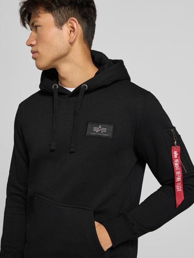 Männermodel trägt Hoodie mit Känguru-Tasche und rückseitigem Label-Print von Alpha Industries, Baumwoll-Mix, Nahaufnahme Oberkörper.