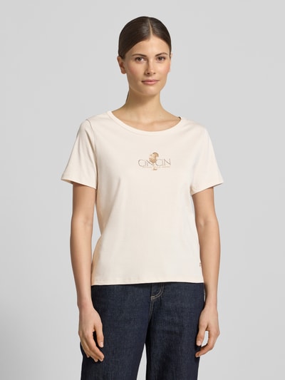 Frauenmodel trägt T-Shirt mit Motiv-Print und Strasssteinbesatz von monari, Reine Baumwolle, Gerader-Schnitt, Rundhalsausschnitt, Motiv mit Strasssteinbesatz, Rückenlänge bei Größe 36 ist 57 cm, dezentes Creme, glatte Textur, Schrift und Motiv in Goldton, Ganzkörperansicht von vorne.